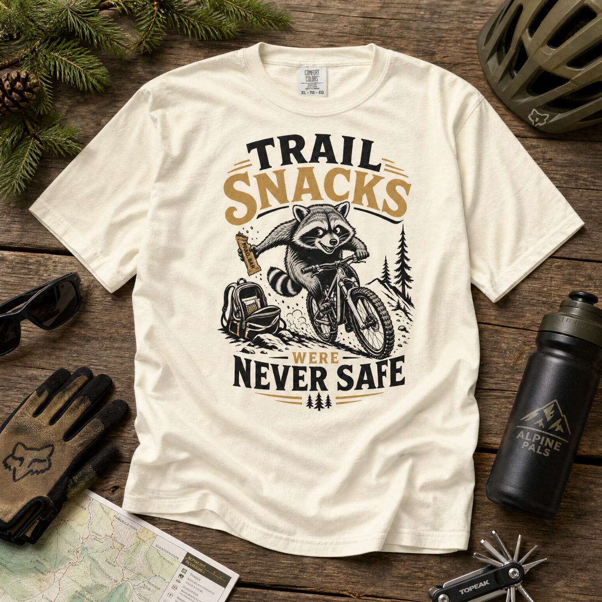 Ivory colored t-shirt with raccoon graphic and 'Trail Snacks Were Never Safe' text on a wooden surface with outdoor gear.