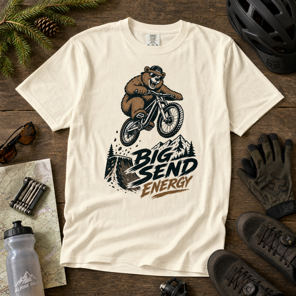 Ivory t-shirt with bear riding a bike graphic and 'Big Send Energy' text on a wooden surface with outdoor gear.