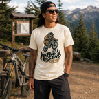 Man wearing a t-shirt with a bear riding a bike graphic and 'Big Send Energy' text, standing in a mountainous area with a bike and trees in the background.