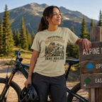 Woman in a 'Professionally Unqualified Trail Builder' t-shirt standing next to a bicycle with mountains in the background.