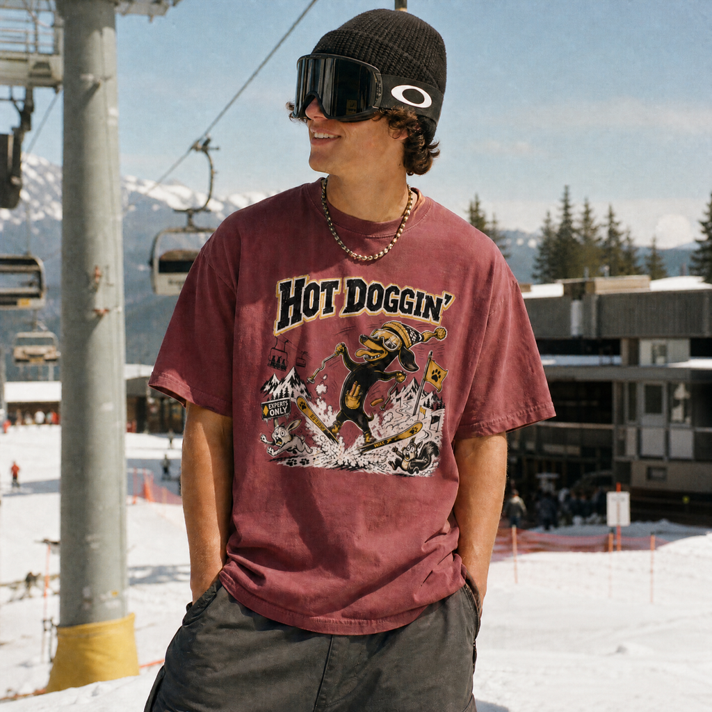 Person wearing a brick colored t-shirt with a graphic design, standing on a snowy mountain with ski lifts and trees in the background.