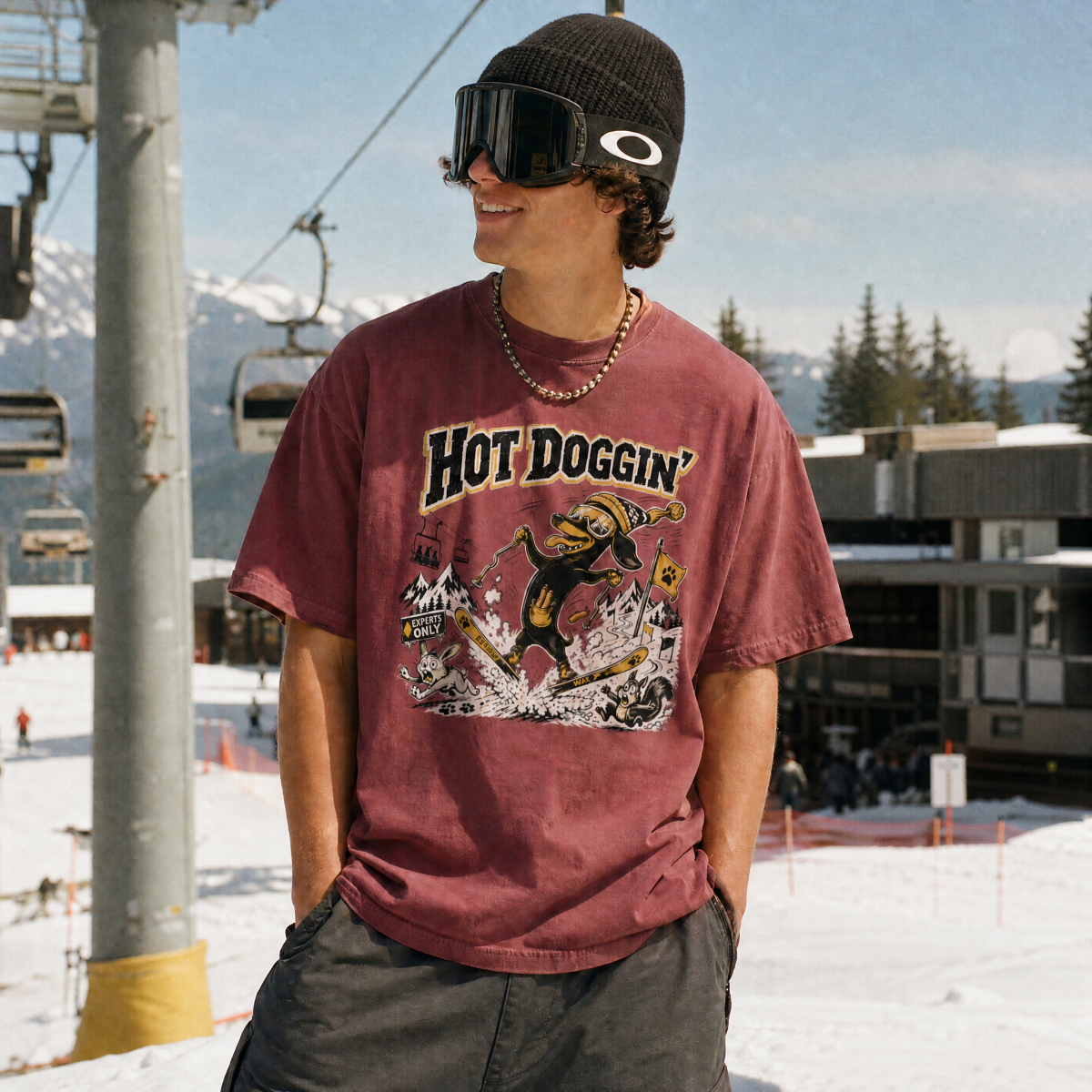 Person wearing a brick colored t-shirt with a graphic design, standing on a snowy mountain with ski lifts and trees in the background.