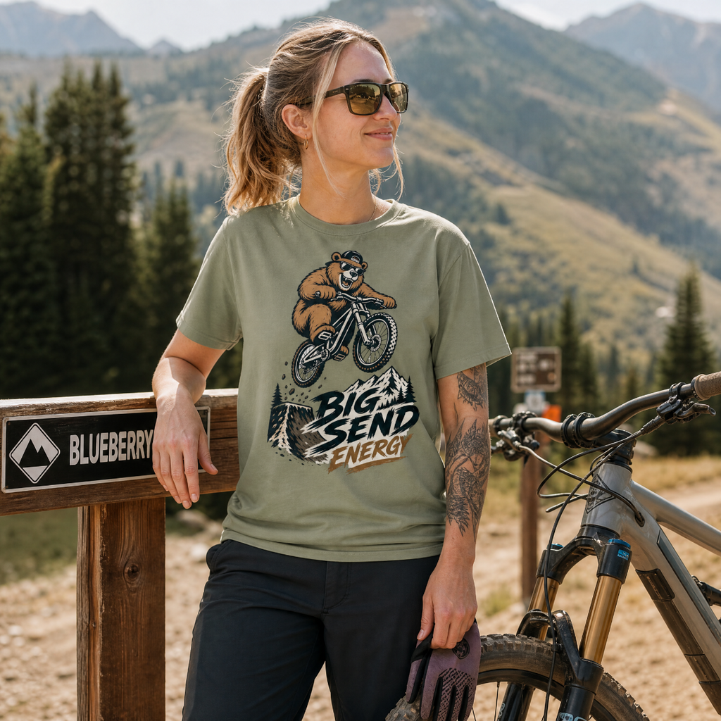 Woman in a mountainous area wearing a t-shirt with a bear graphic and text, standing next to a bike.
