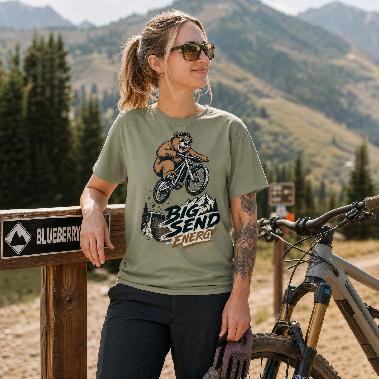 Woman in a mountainous area wearing a t-shirt with a bear graphic and text, standing next to a bike.