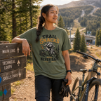 Person wearing a 'Trail Snacks Never Safe' t-shirt in a mountainous area with a bike and signpost.