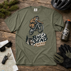 Moss green t-shirt with bear riding a bike graphic and 'Big Send Energy' text on a wooden surface with accessories.