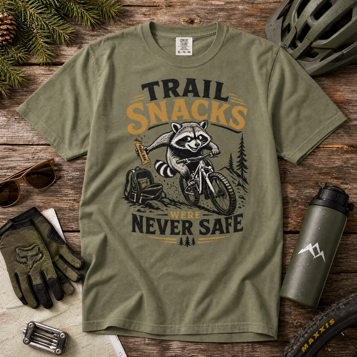 Moss green t-shirt with a raccoon graphic and text on a wooden surface with outdoor accessories.