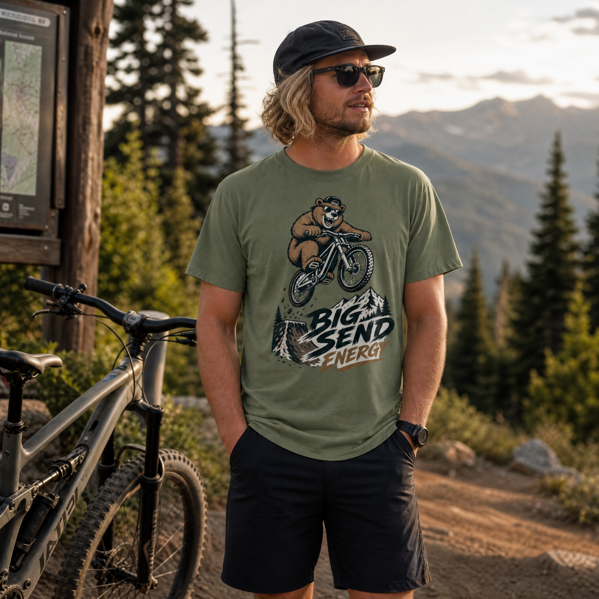 Man wearing a t-shirt with a bear graphic and 'Big Send Energy' text, standing next to a bike in a mountainous area.