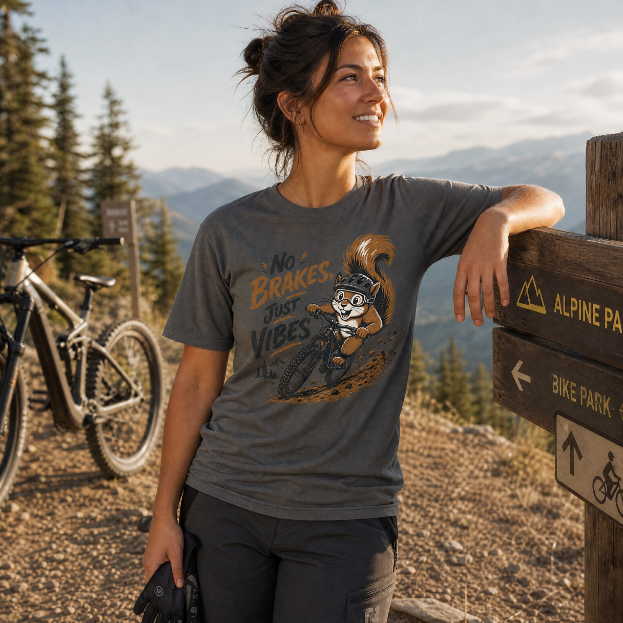 Woman in a mountain biking shirt with a squirrel graphic, standing next to a bike in a forested area.