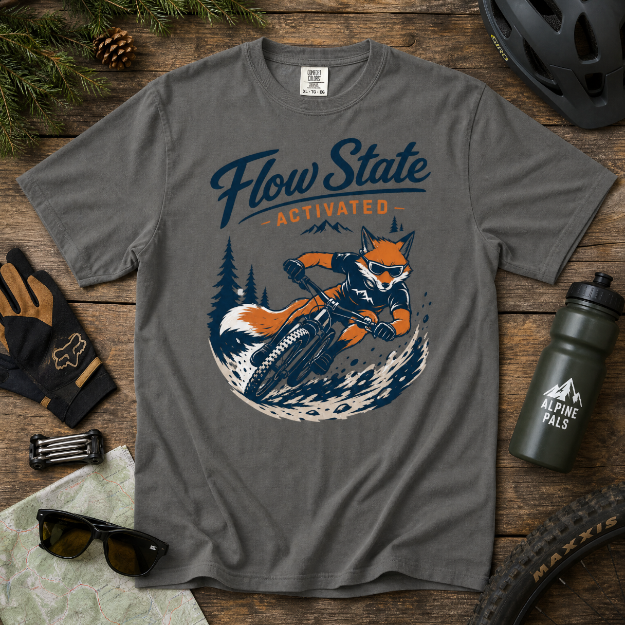 Gray t-shirt with a fox riding a bike graphic and 'Flow State Activated' text on a wooden surface with outdoor gear.