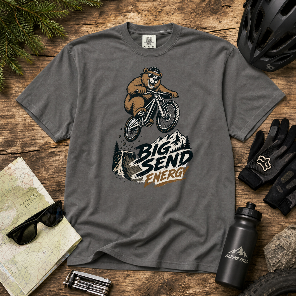 Pepper gray t-shirt with bear riding a bike graphic and 'Big Send Energy' text on a wooden surface with accessories.