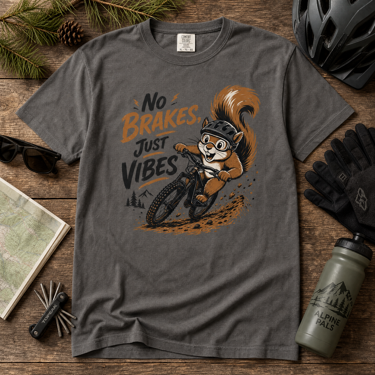 Gray t-shirt with a cartoon squirrel on a bike and text 'No Brakes, Just Vibes' on a wooden surface with cycling accessories.